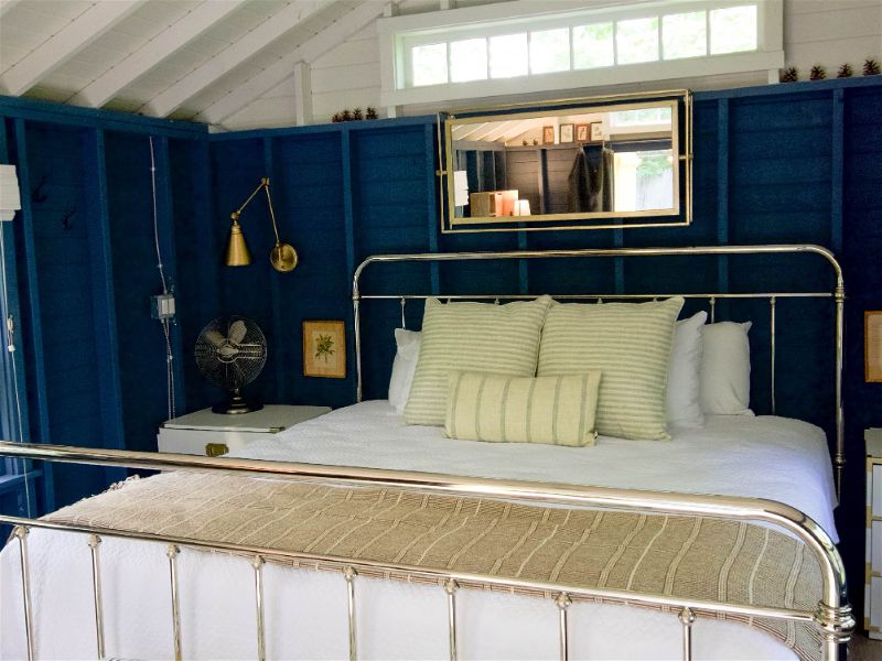 Quarto com paredes de madeira pintadas de azul escuro e cama de ferro com tecido e travesseiros com cores claras e listradas