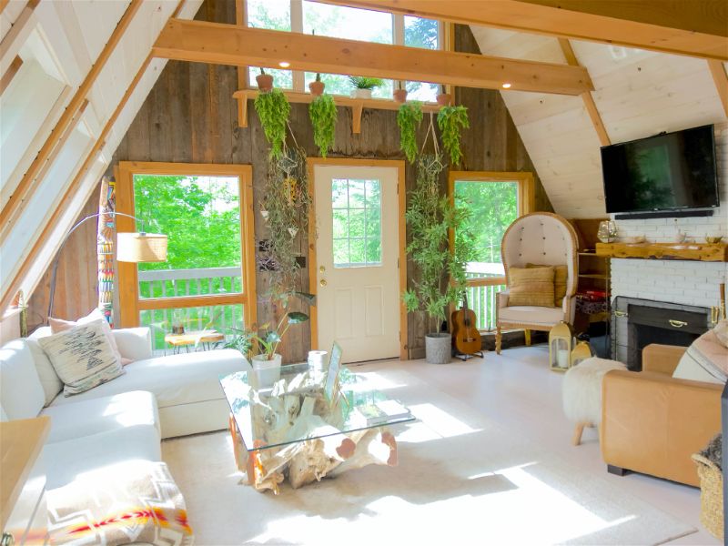 Sala com decoração estilo boho, estrutura da casa em madeira, móveis claros e alguns tecidos coloridos e algumas plantas penduradas na parede próximo porta