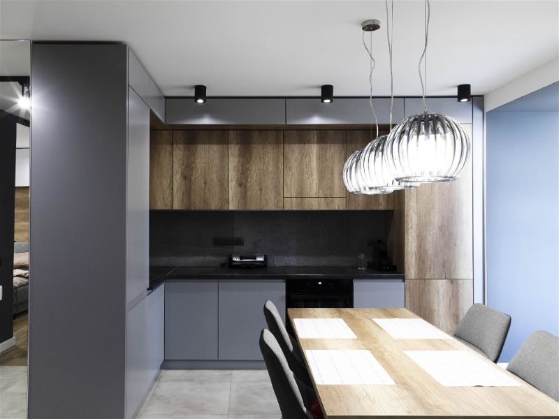 Cozinha moderna e pequena com tons de madeira e cinza, com iluminação sobre mesa de jantar, bem como, por spots espalhados ao redor da cozinha